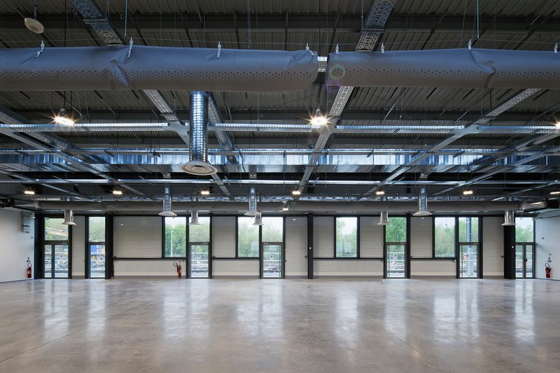Open-plan interior space with exposed ceiling ducts, pendant lights, and a glazed facade overlooking greenery