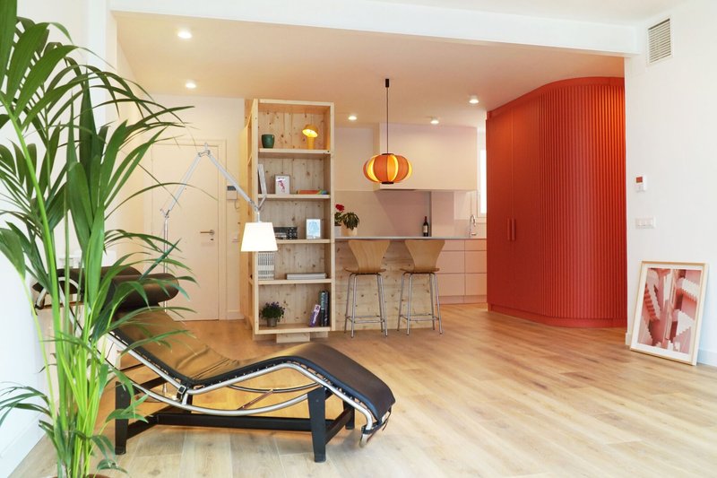 Open-plan living space with light wood flooring, red ribbed storage volume and plywood shelving unit