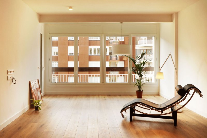 Pale interior room with wide window overlooking residential buildings and a lounge chair on timber floor