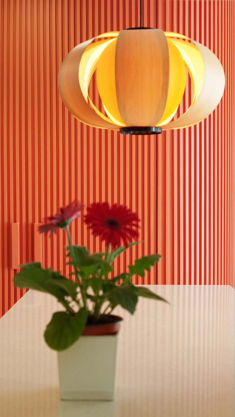 Pendant lamp with layered wood veneer shade above a potted gerbera against red vertical slats