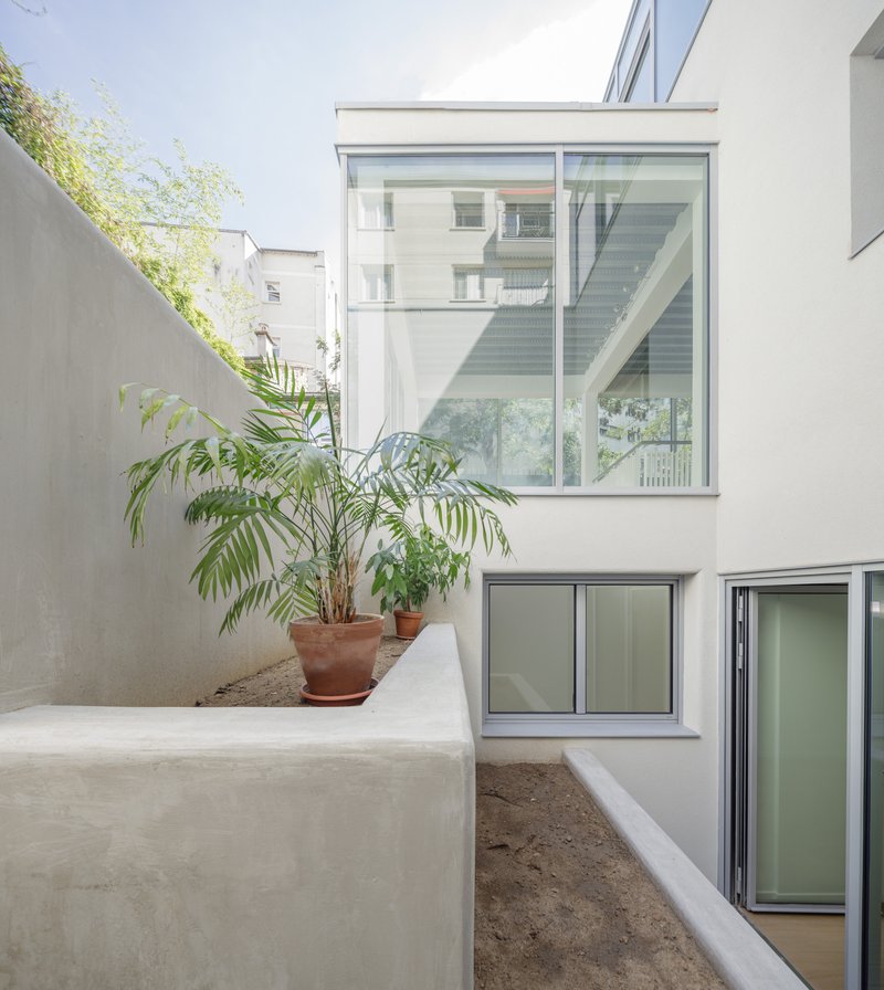 Courtyard view showing stacked glazed volumes with rendered planter bed and potted palms against neighboring concrete wall