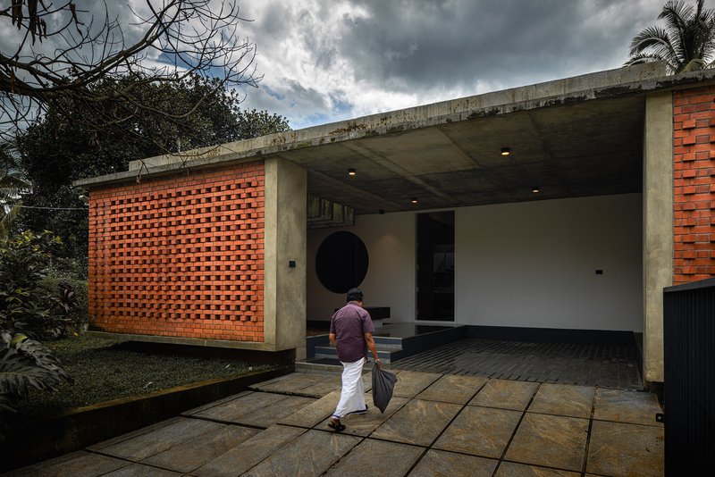 Covered entrance with perforated brick screen and concrete overhang as a person walks across stone paving under cloudy skies