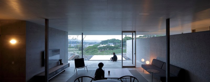 Living room interior at dusk with two figures seated and terrace opening to hillside beyond