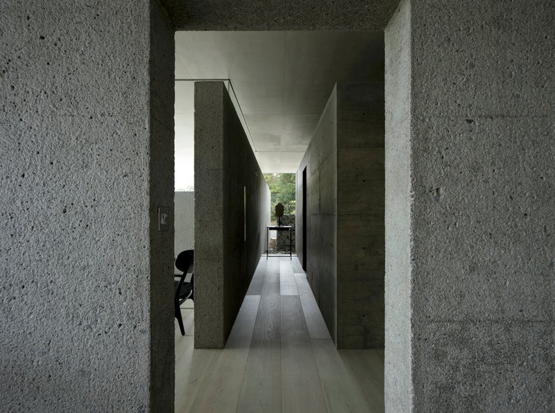 Corridor view through textured concrete walls leading to glazed opening with figure at table