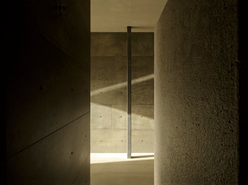 Narrow hallway in board-formed concrete with vertical slot window casting sunlight on textured stone wall