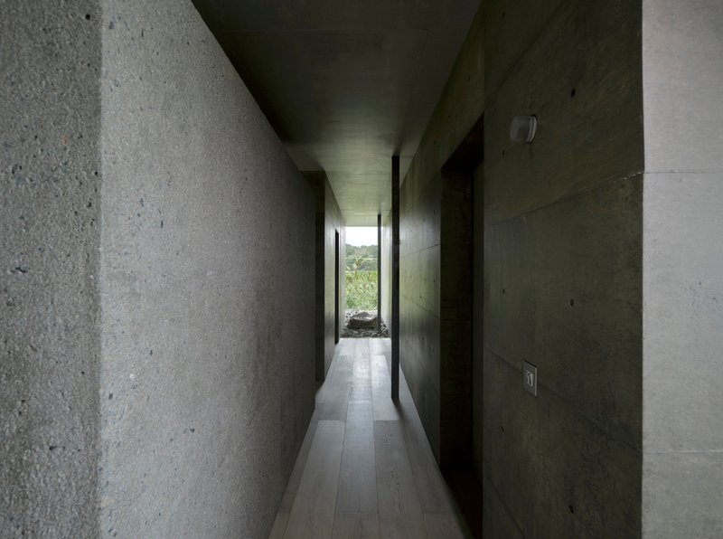 Long corridor lined with exposed concrete and rough stone walls leading to a sliver of garden