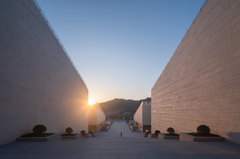 Paved courtyard framed by two angled brick-clad walls converging toward a distant hillside at sunset