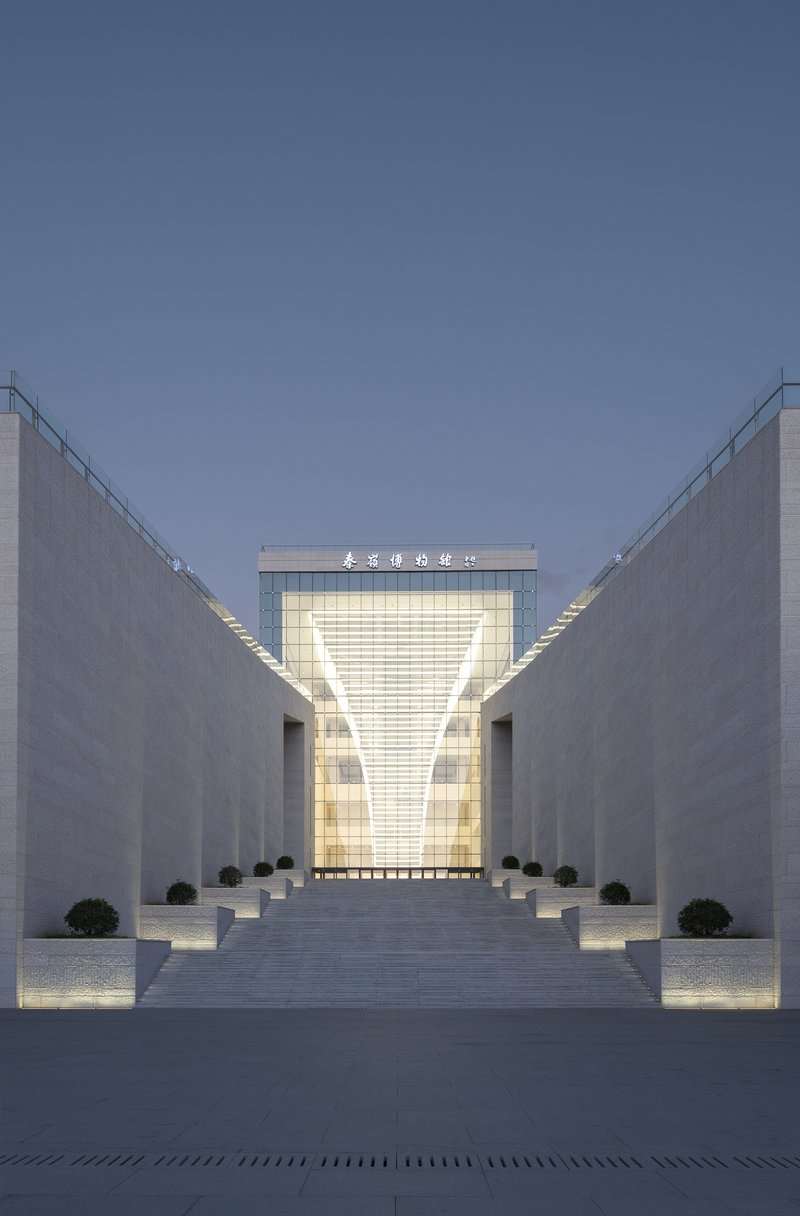 Stone-paved plaza flanked by angled walls leading to the illuminated glass entrance at dusk