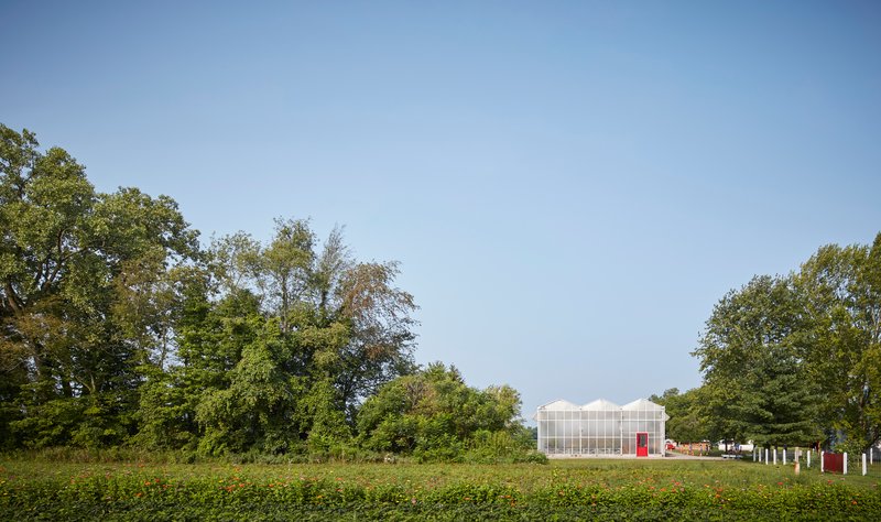 Greenhouse pavilion with translucent polycarbonate walls and red doors viewed across a green field beneath mature trees