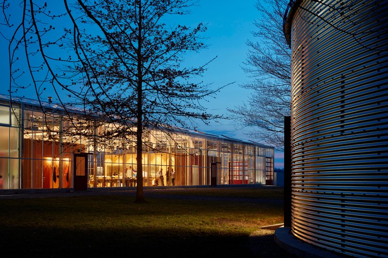 Glass-walled pavilion glowing from interior lights at dusk with bare trees and a cylindrical metal form nearby