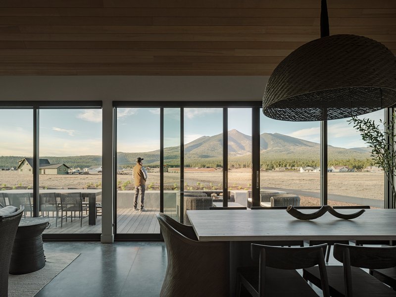 Dining area with full-height glazing and a figure standing on the terrace framed by distant peaks