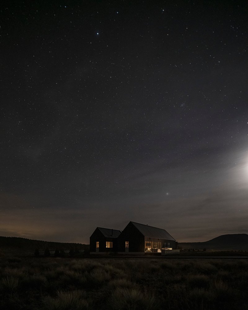 Twin gabled volumes illuminated from within across a scrubland field under a star-filled night sky