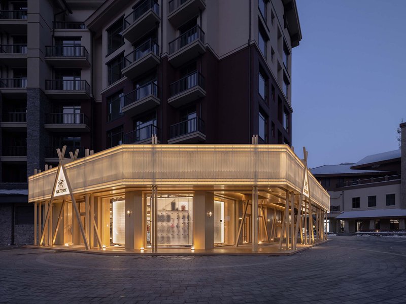 Illuminated corner storefront with diagonal timber columns and translucent panels at dusk