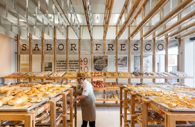 Baker walking past tiered wooden display shelves beneath a timber frame and translucent ceiling panels