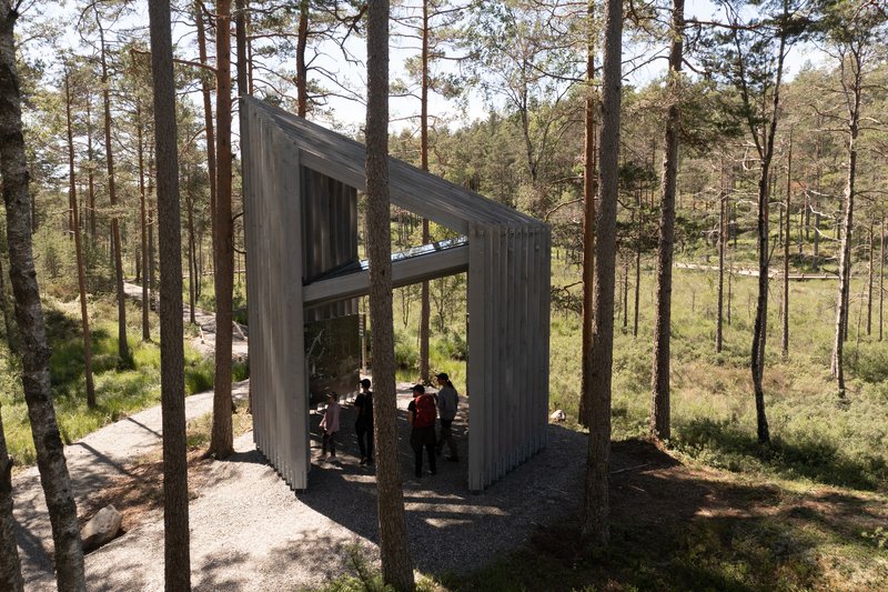 Open-air pavilion with vertical timber screens framing visitors seated beneath an angled roof plane
