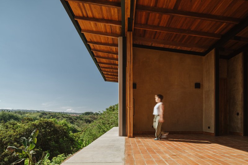 Covered terrace with exposed timber ceiling, terracotta tile floor, and figure walking past rammed earth wall