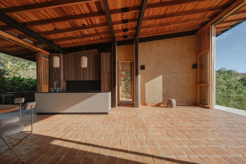 Open plan room with terracotta tile floor, rammed earth walls, and timber slatted door receiving afternoon sun