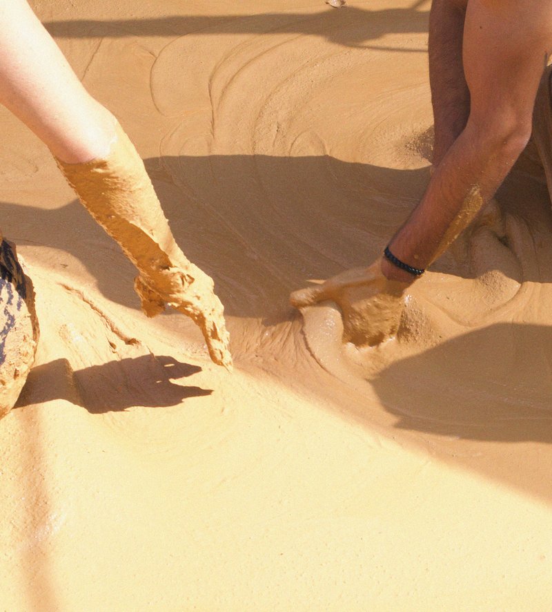 Hands smoothing earthen plaster onto curved floor surface in bright sunlight