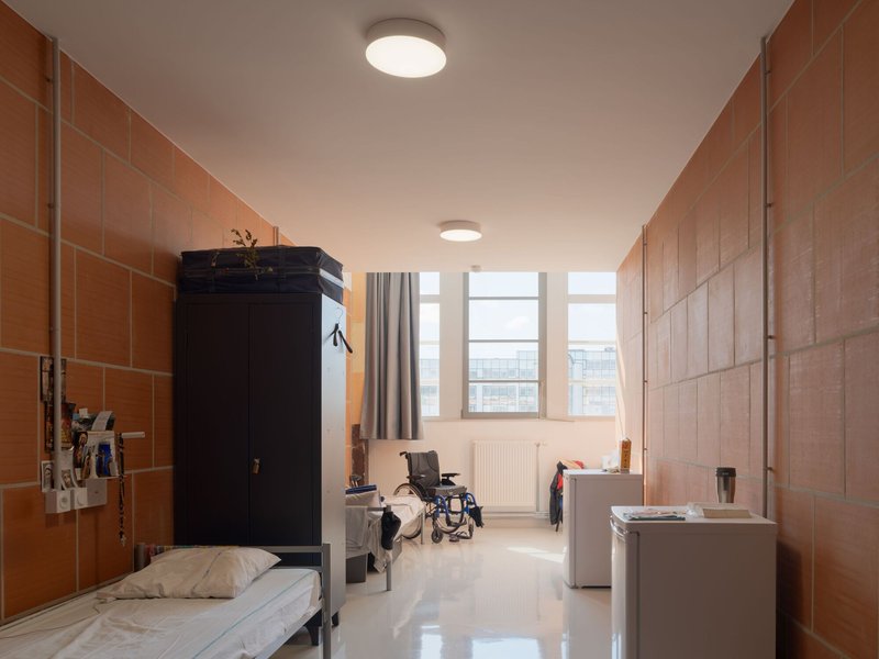 Residential room with terracotta tile walls, metal bed, black wardrobe, and wheelchair visible by window