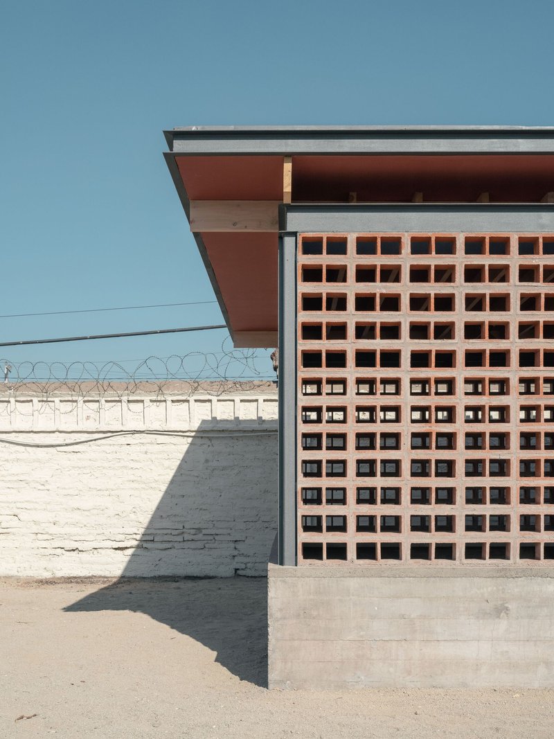 Perforated terracotta block screen beneath deep overhangs with barbed wire perimeter wall beyond