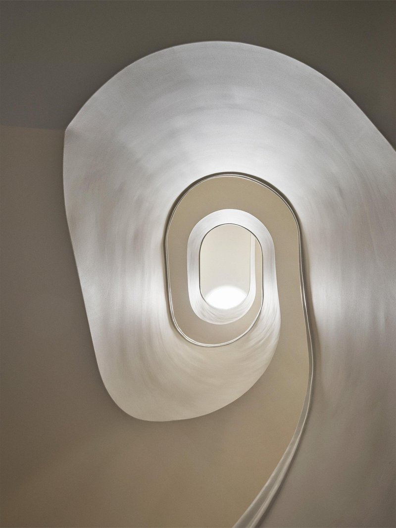 Looking up through a spiral stairwell with oval openings and smooth plaster walls in soft daylight