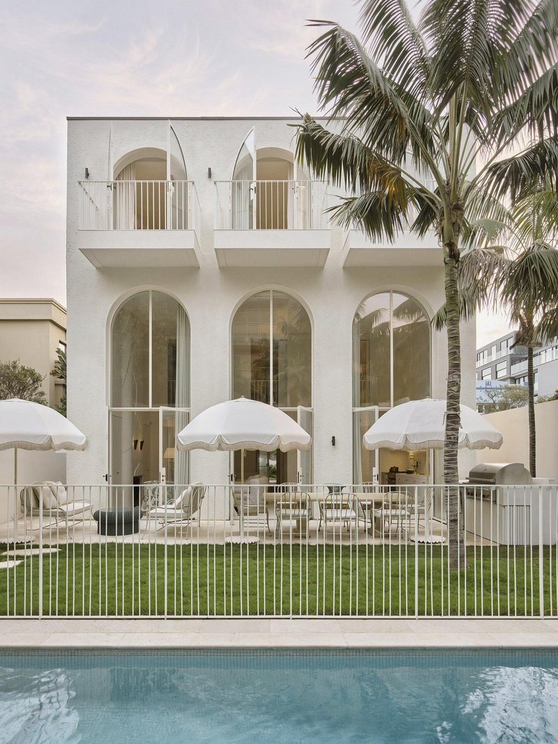 White stucco facade with arched openings and upper balconies beside a pool and palm trees