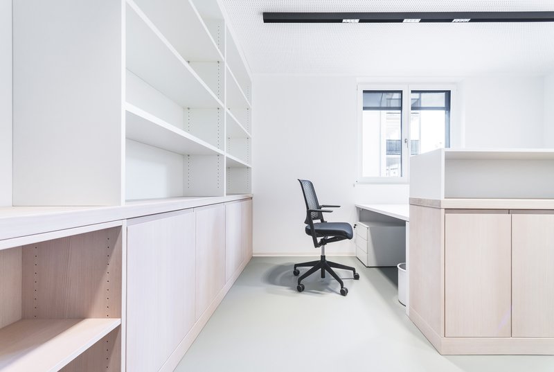 Individual workstation with white cabinetry and mesh office chair in narrow workspace with grey flooring