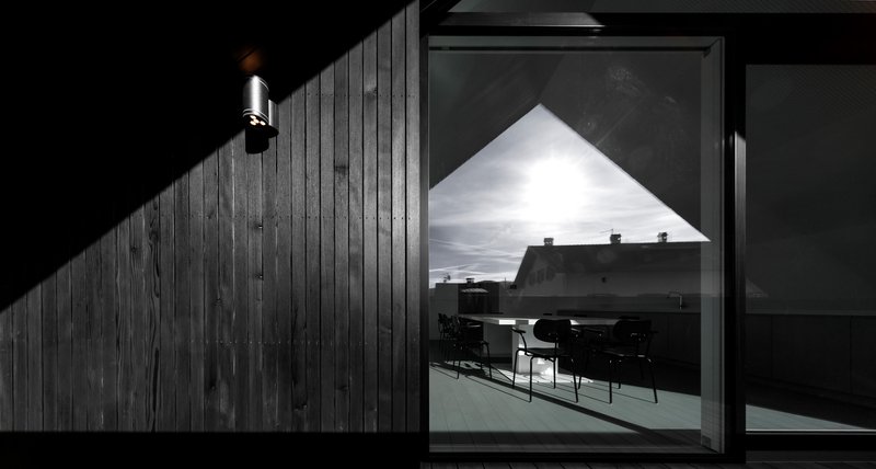 Triangular window reflecting interior dining area at night with exterior sconce illuminating charred timber cladding