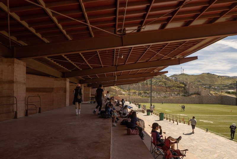 Open-air concourse with exposed timber joists and spectators seated on benches overlooking the field