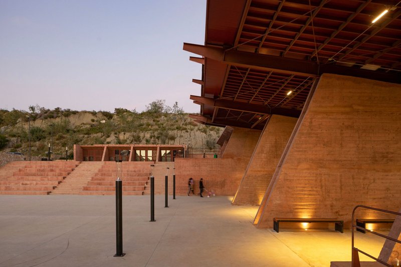 Cantilevered timber-framed canopy over a concrete plaza with stepped seating at dusk
