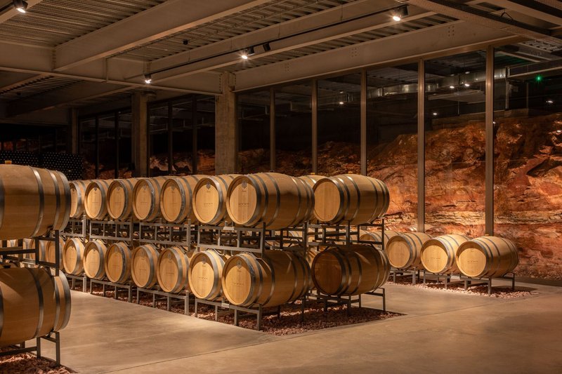 Barrel room with wooden wine casks on metal racks against an exposed red rock wall