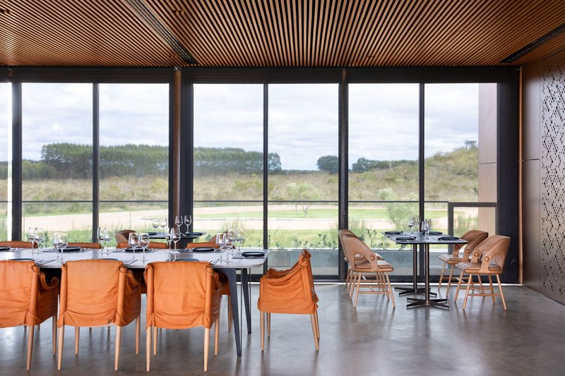 Dining space with orange upholstered chairs and slatted timber ceiling facing expansive glazing and landscape beyond