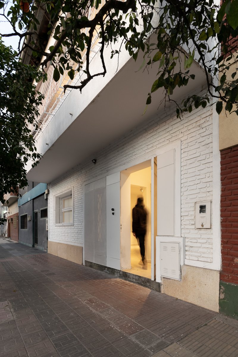 White painted brick facade with open door revealing illuminated interior at dusk with a blurred figure