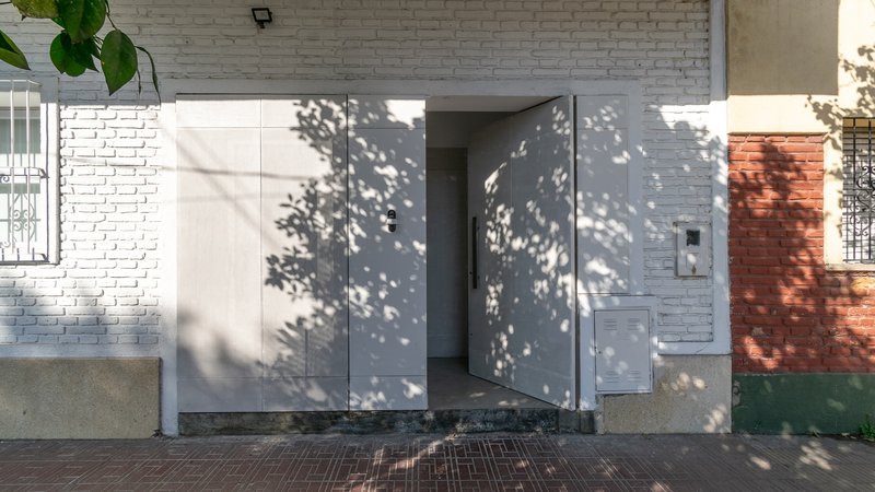 Street facade with pivoting white steel doors partially open, casting dappled tree shadows across the brick wall