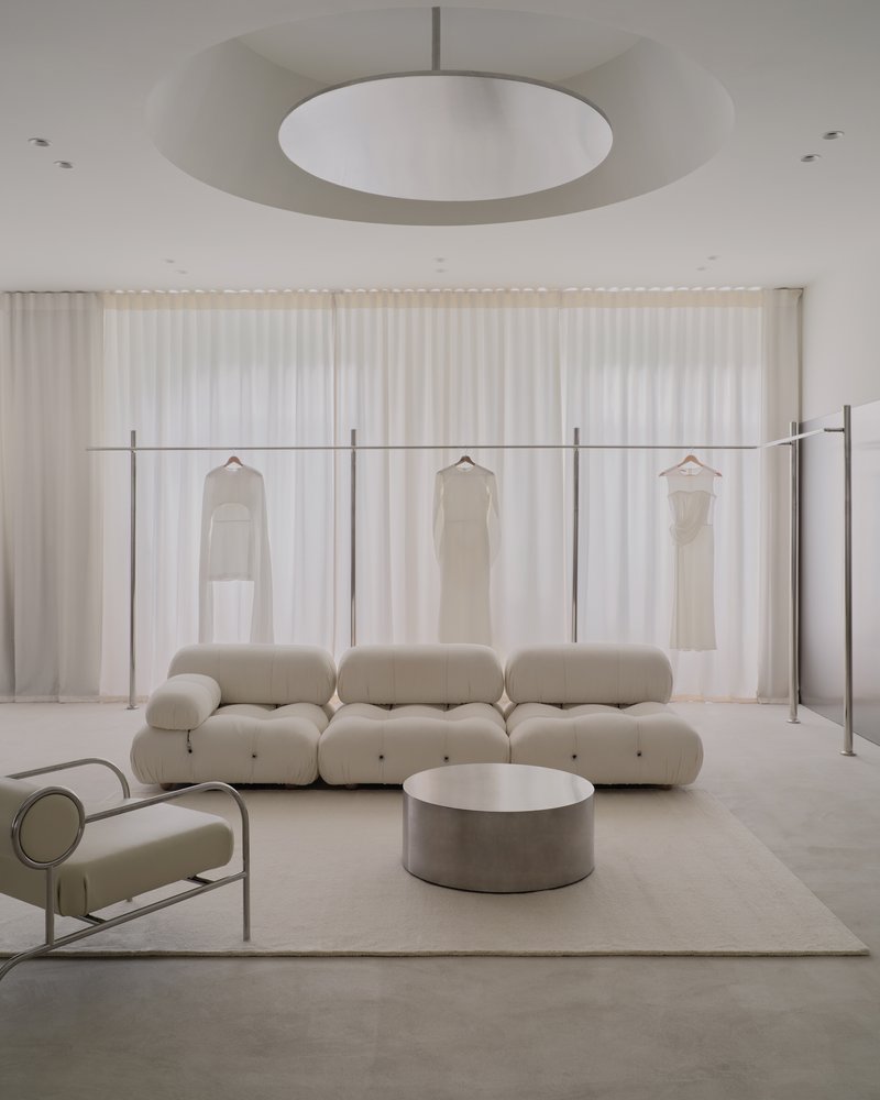 Freestanding steel clothing rack with hanging garments against sheer curtains and a disc pendant light