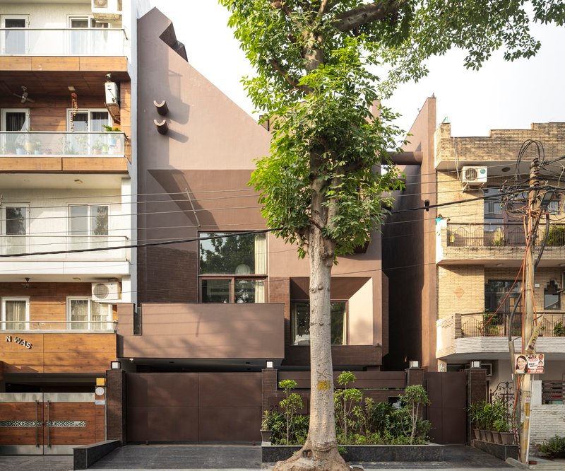 Street facade with layered brown and beige panels framed by a mature tree and neighboring buildings