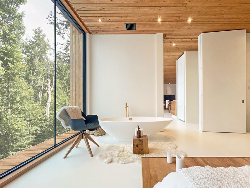 Freestanding bathtub on white flooring beneath timber ceiling with full-height glazing to forest beyond
