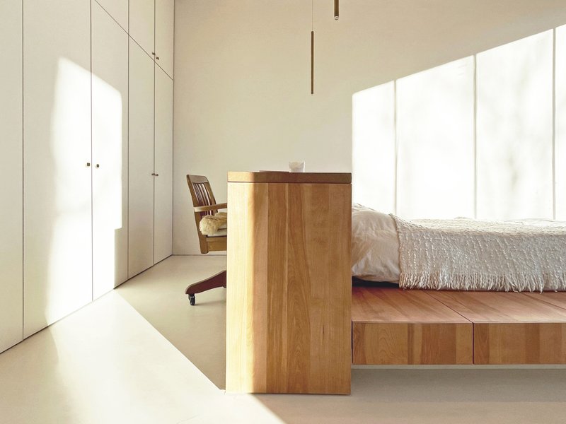 Minimalist bedroom with timber platform bed and floor-to-ceiling white storage cabinetry bathed in natural light