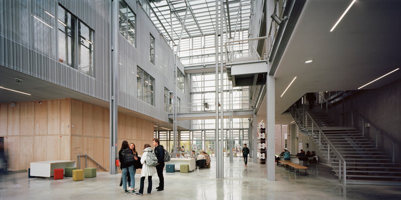 Multi-level lobby with glass roof, metal staircase, and groups of visitors gathered near wood paneled walls