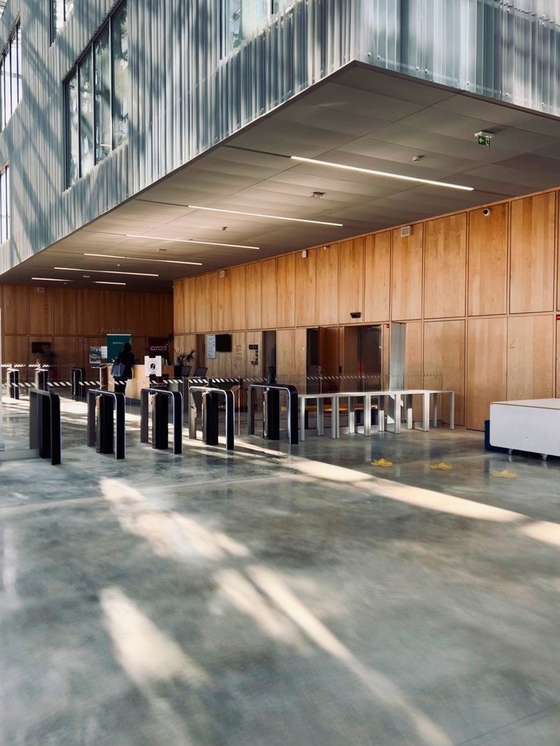 Entry lobby showing timber wall panels and turnstile gates beneath a concrete soffit with linear light fixtures