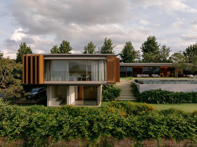 Two-story volume with vertical timber slats and full-height glazing overlooking a garden and pool under cloudy skies