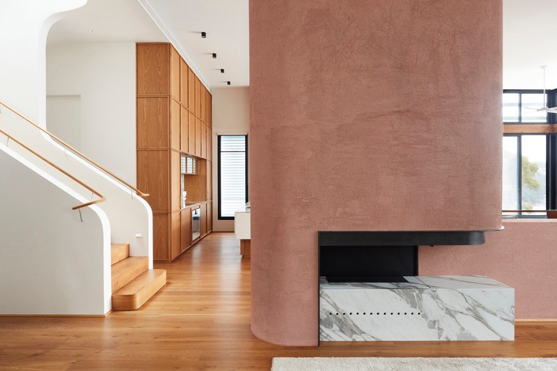Cylindrical rammed earth fireplace with marble hearth beside timber cabinetry and curved stair