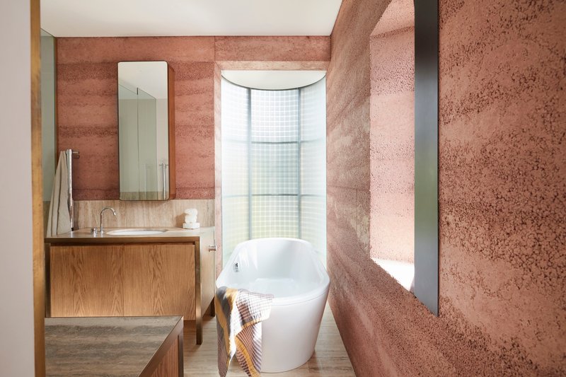 Bathroom with rammed earth walls, freestanding bathtub and curved window framing daylight