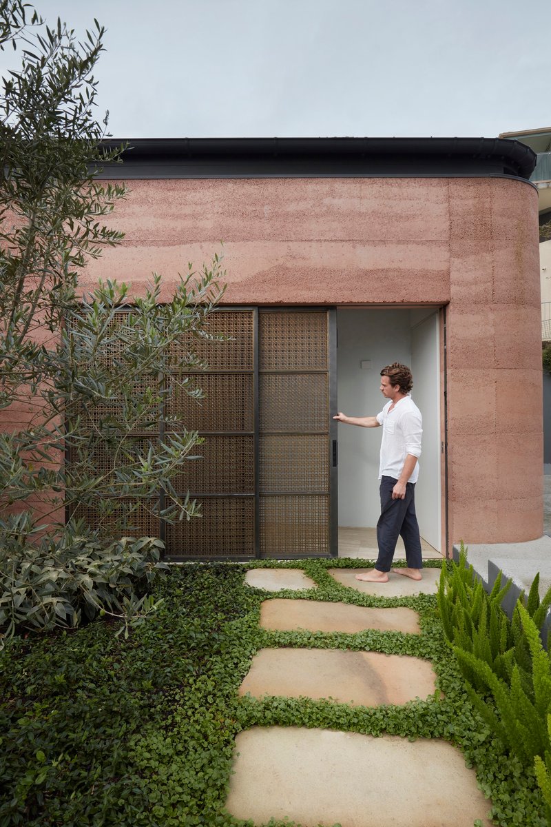 Rammed earth entry facade with perforated metal sliding door and stepping stones through ground cover planting