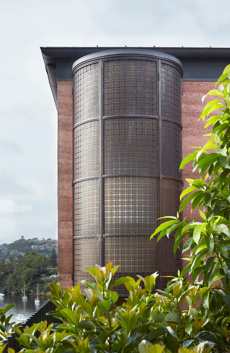 Curved perforated metal screen tower rising against rammed earth wall with hillside visible beyond vegetation