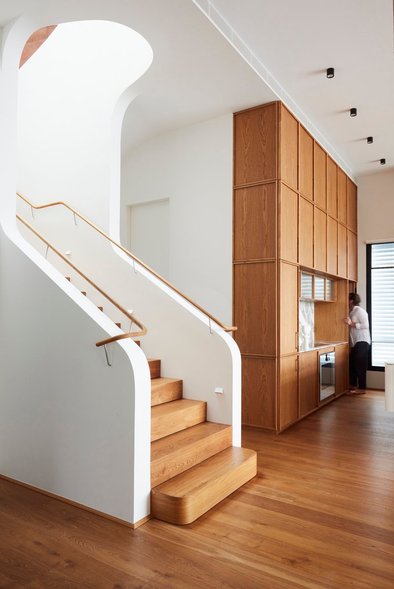 Timber staircase with brass handrail ascending beside full-height oak cabinetry wall with integrated appliances