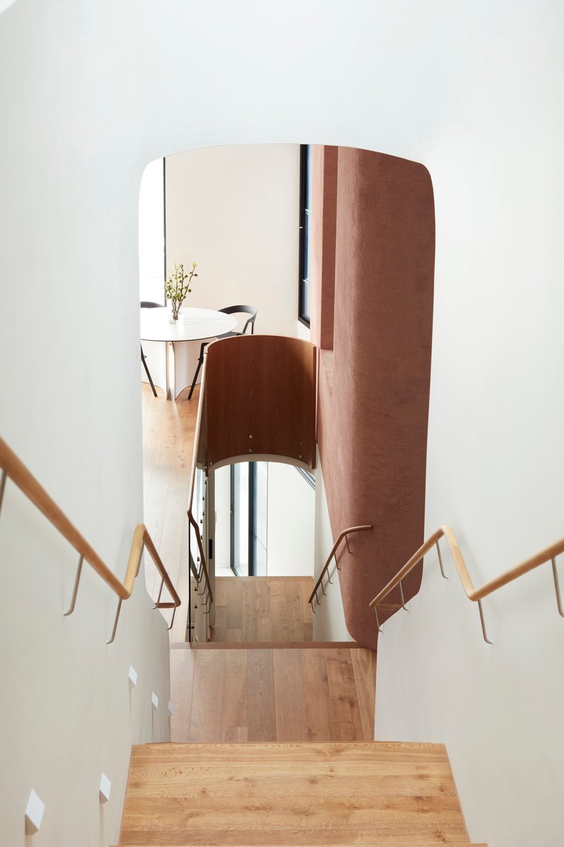 Downward view of timber staircase with curved terracotta-clad walls and rounded mirror at the landing