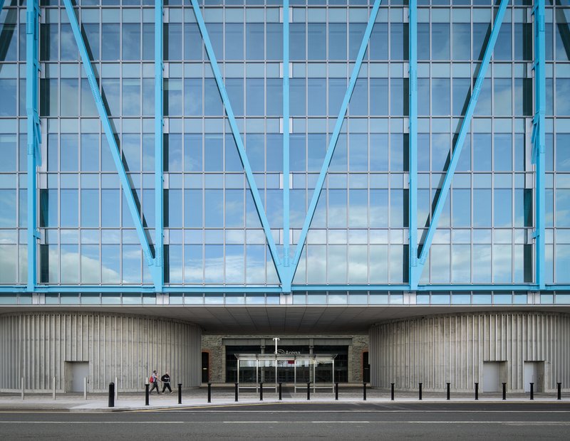 Facade detail showing blue diagonal steel bracing over a glazed curtain wall with pedestrians passing below