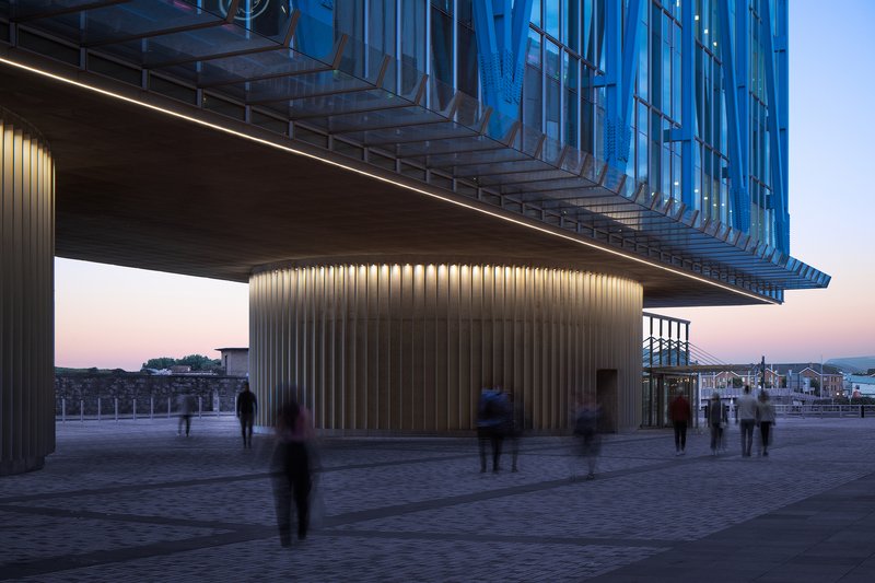 Illuminated ribbed concrete base volume supporting a cantilevered glass tower at dusk with pedestrians crossing the plaza