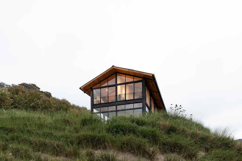 Two-story gabled facade with steel-framed glazing and timber cladding atop a grassy dune